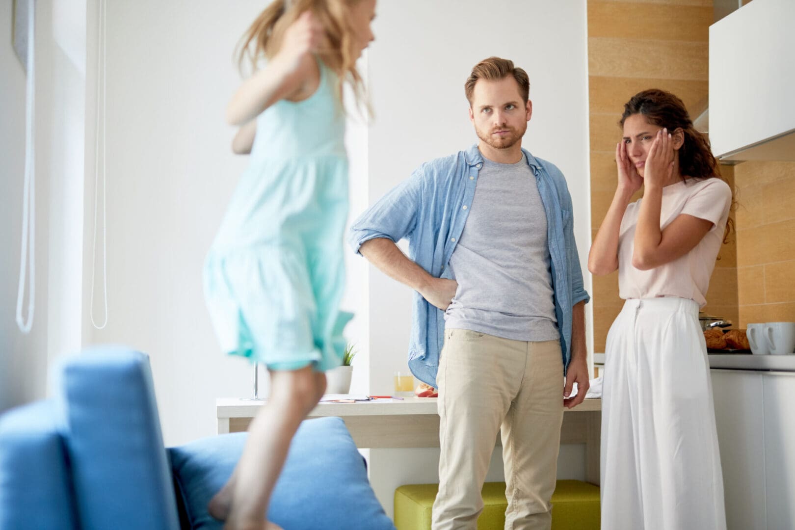 Shocked father and mother looking at their little naughty daughter jumping on armchair in the kitchen